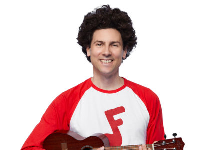 Jeremy Fisher, a white man with curly brown hair, grins at the camera. He is holding a ukulele and wearing a red and white shirt with a red Jeremy and Jazzy logo