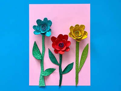 Three cardboard painted flowers on a pink card against a blue background.